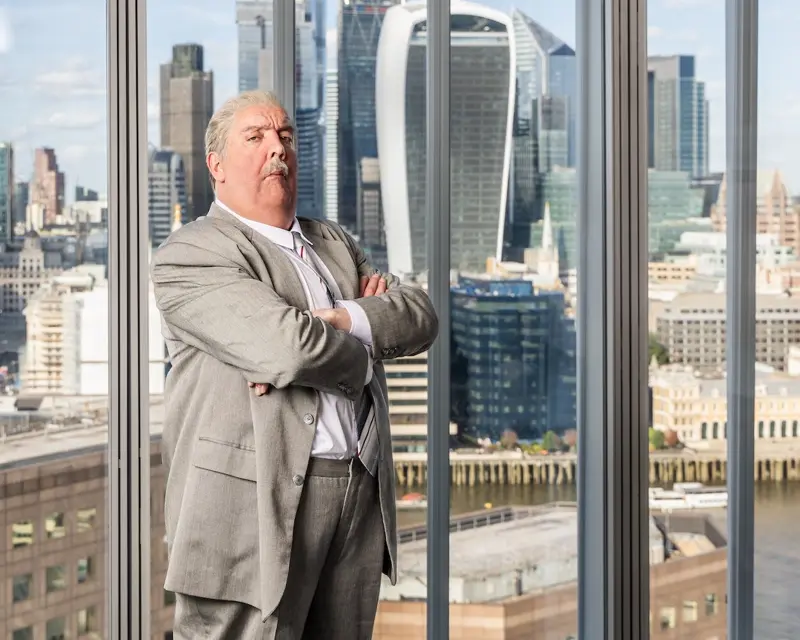 Brian Butterfield, arms folded, in front of a floor-to-ceiling window overlooking the City of London